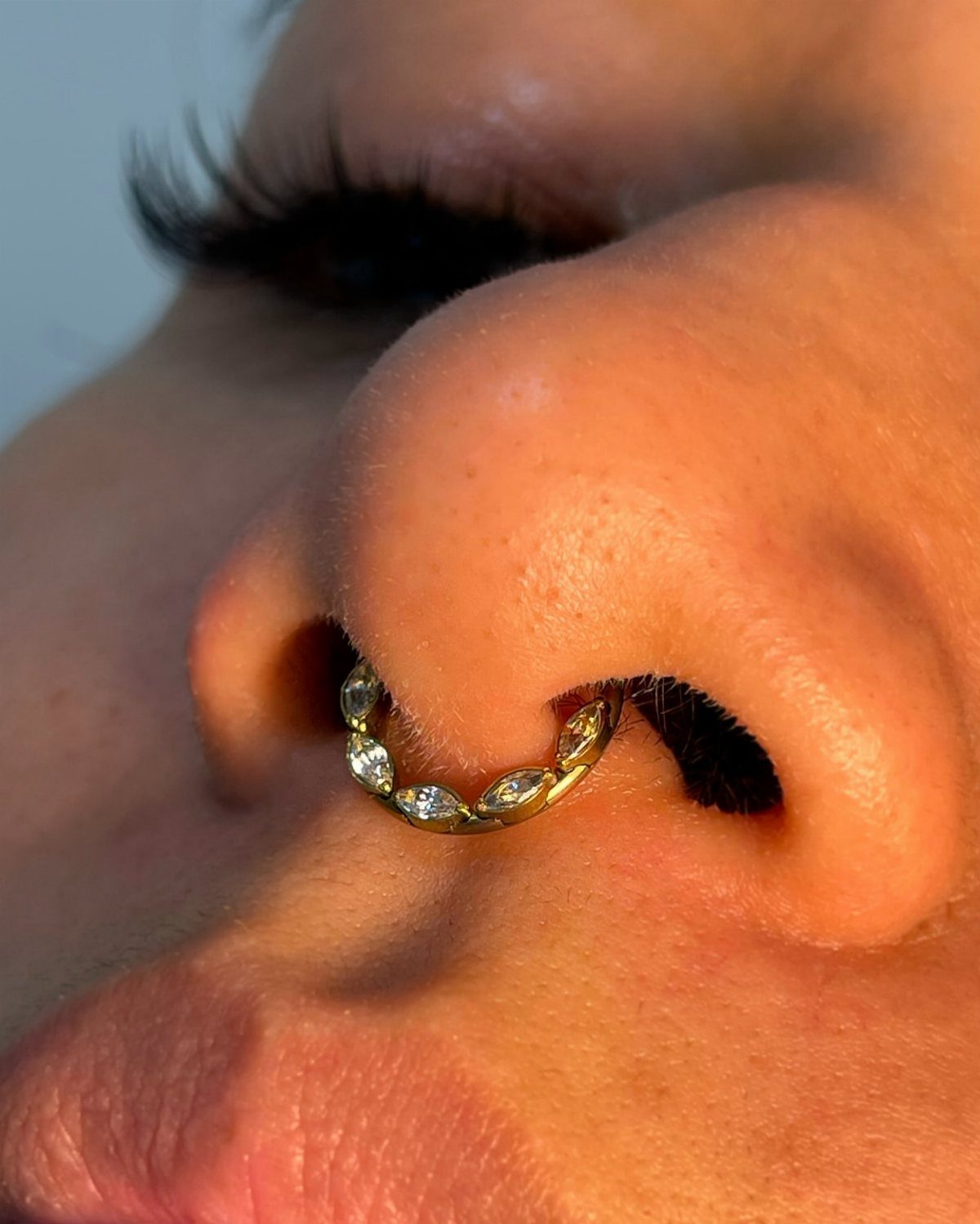 Gold-colored thick septum ring with clear gemstones piercing a person's nasal septum.