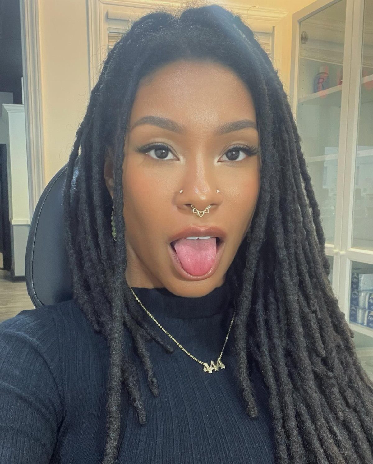 Woman with dreadlocks wearing a gold wing-shaped septum piercing and gold necklace.