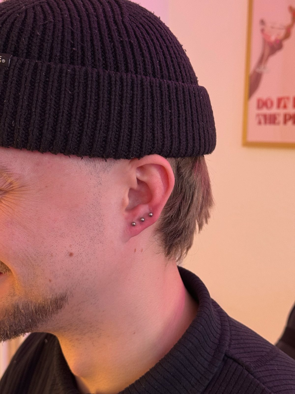 Person's ear with three silver stud piercings in the lobe wearing a black beanie