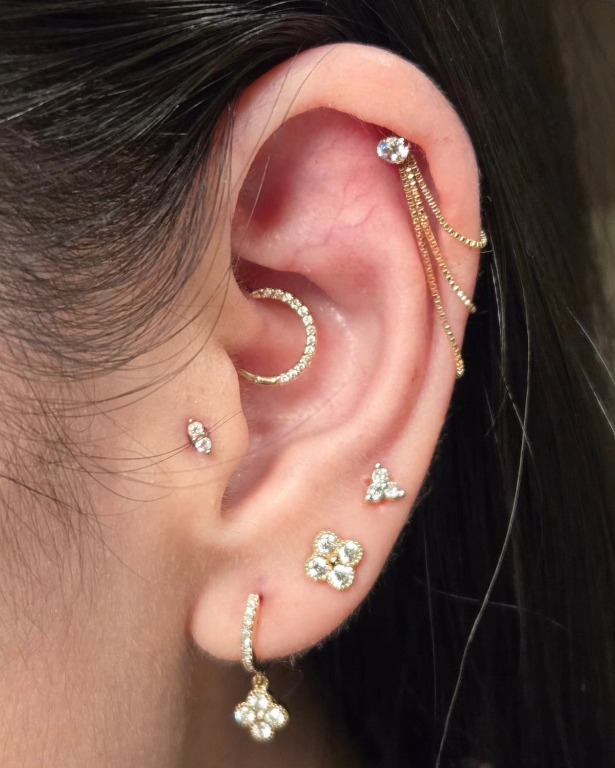 Ear with multiple gold and diamond piercings including floral studs and a helix chain