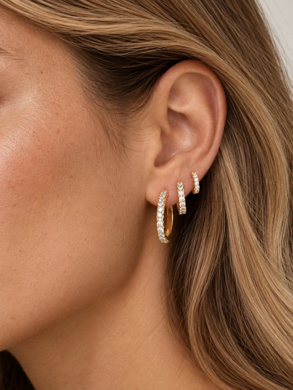Three gold hoop earrings with clear crystals piercing a person's earlobe.
