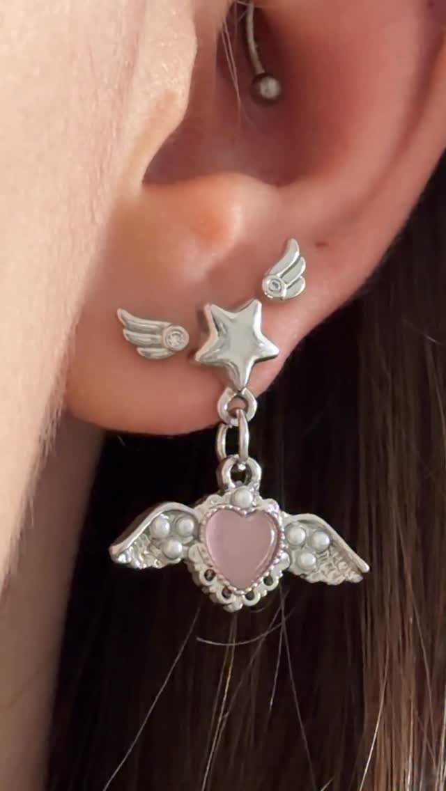 Ear with three silver piercings featuring wing studs and a dangling pink heart charm.