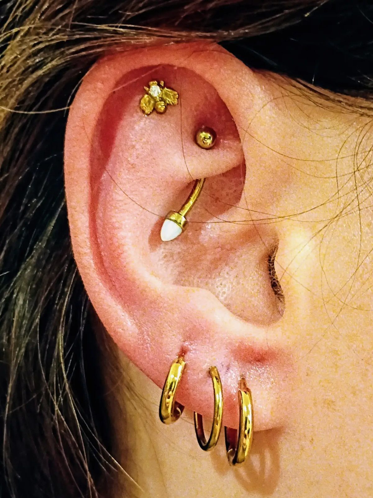 9 Upper Cartilage Piercing Ideas, Jewelry, and Aftercare 8 Human ear with gold upper cartilage piercings, a gold bar, and lobe hoops.