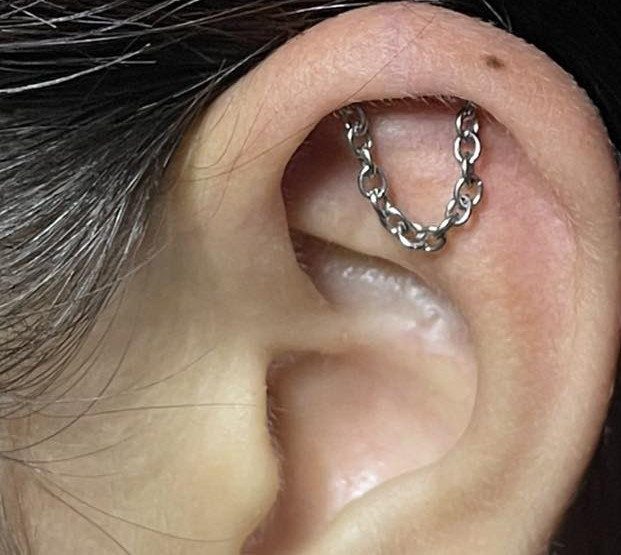 9 Upper Cartilage Piercing Ideas, Jewelry, and Aftercare 4 Human ear with two upper cartilage piercings connected by a silver chain.