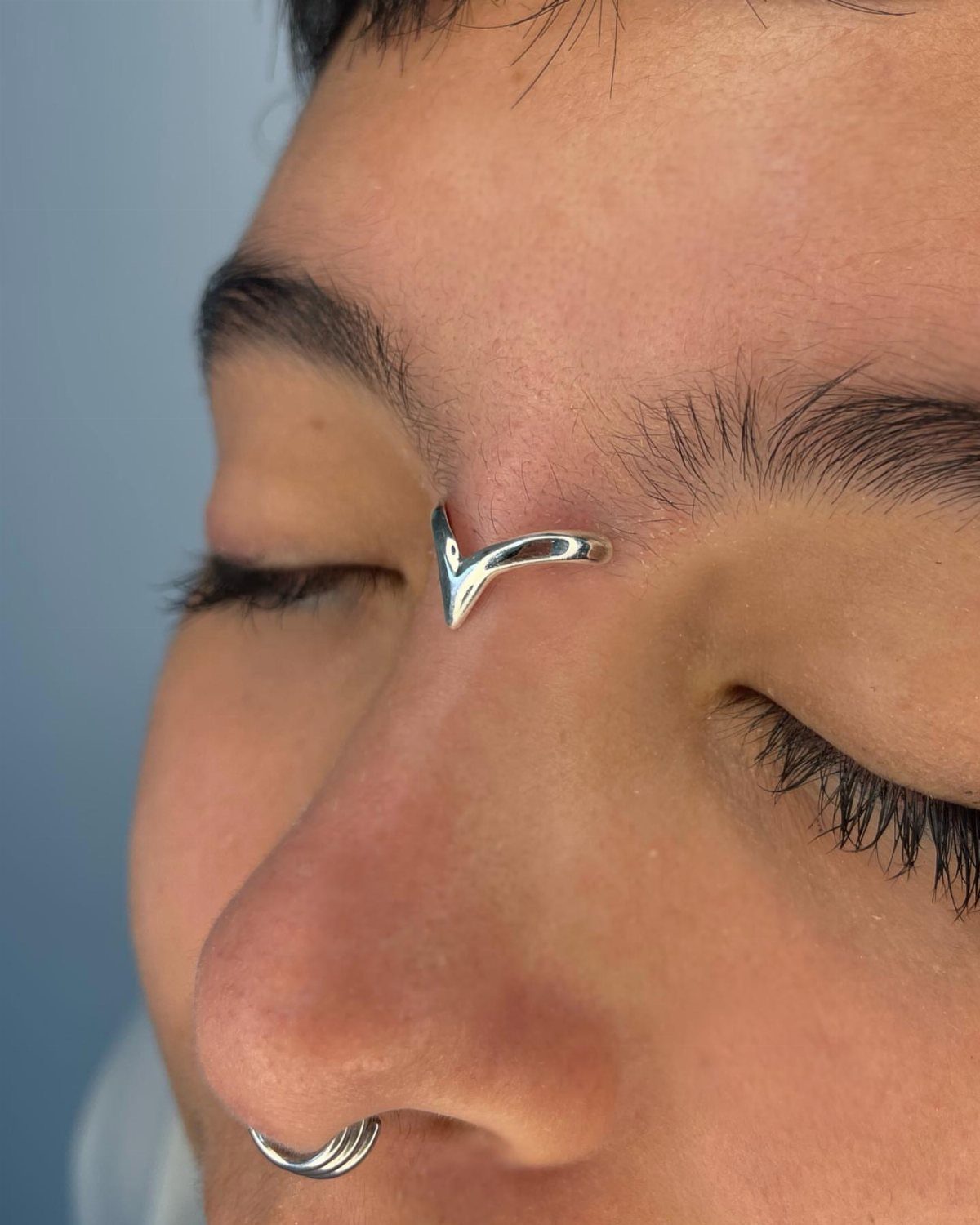 Vertical Bridge Piercing: 7 Jewelry Ideas and Styles 4 Person with a silver V-shaped bridge piercing and a nostril hoop piercing.