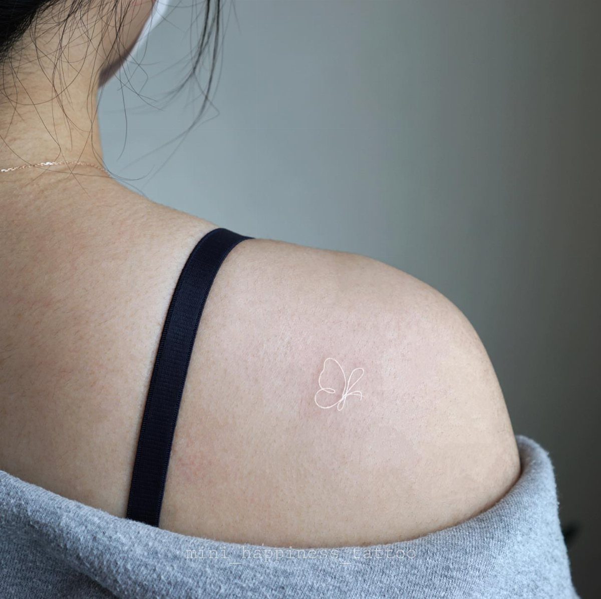 Small white ink butterfly tattoo on a person's shoulder with a minimalist outline.