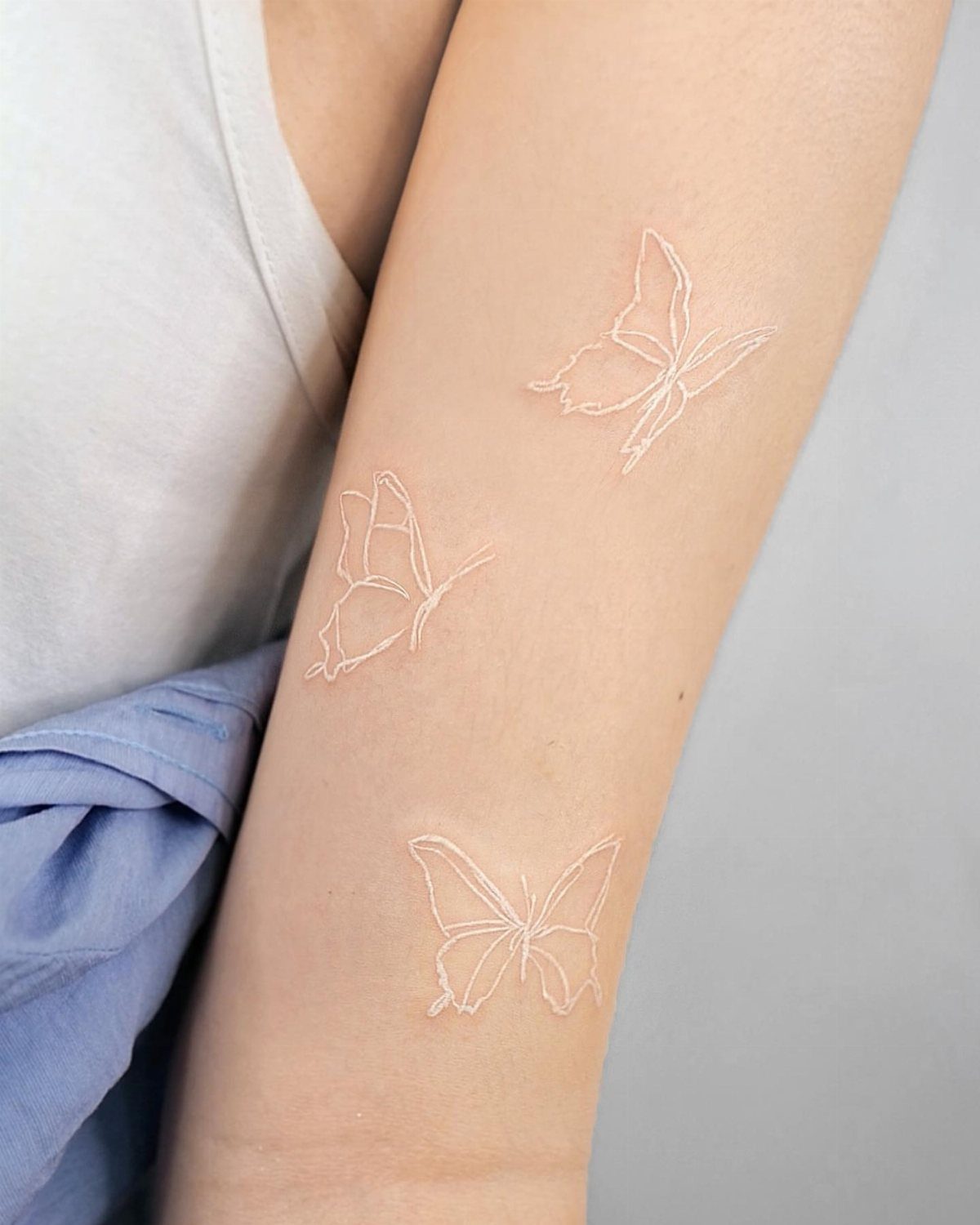 Three white ink butterfly tattoos on a person's arm in a minimalist style